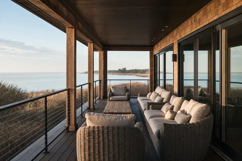 A wide covered porch with dark decking, a cable rail and woven seating for about eight, looking over dune grass to calm water (example photo)