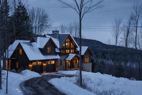 A timber and stone chalet lit at dusk, snow on the ground, bare hardwoods around it and a low wooded ridge above open water behind (example photo)