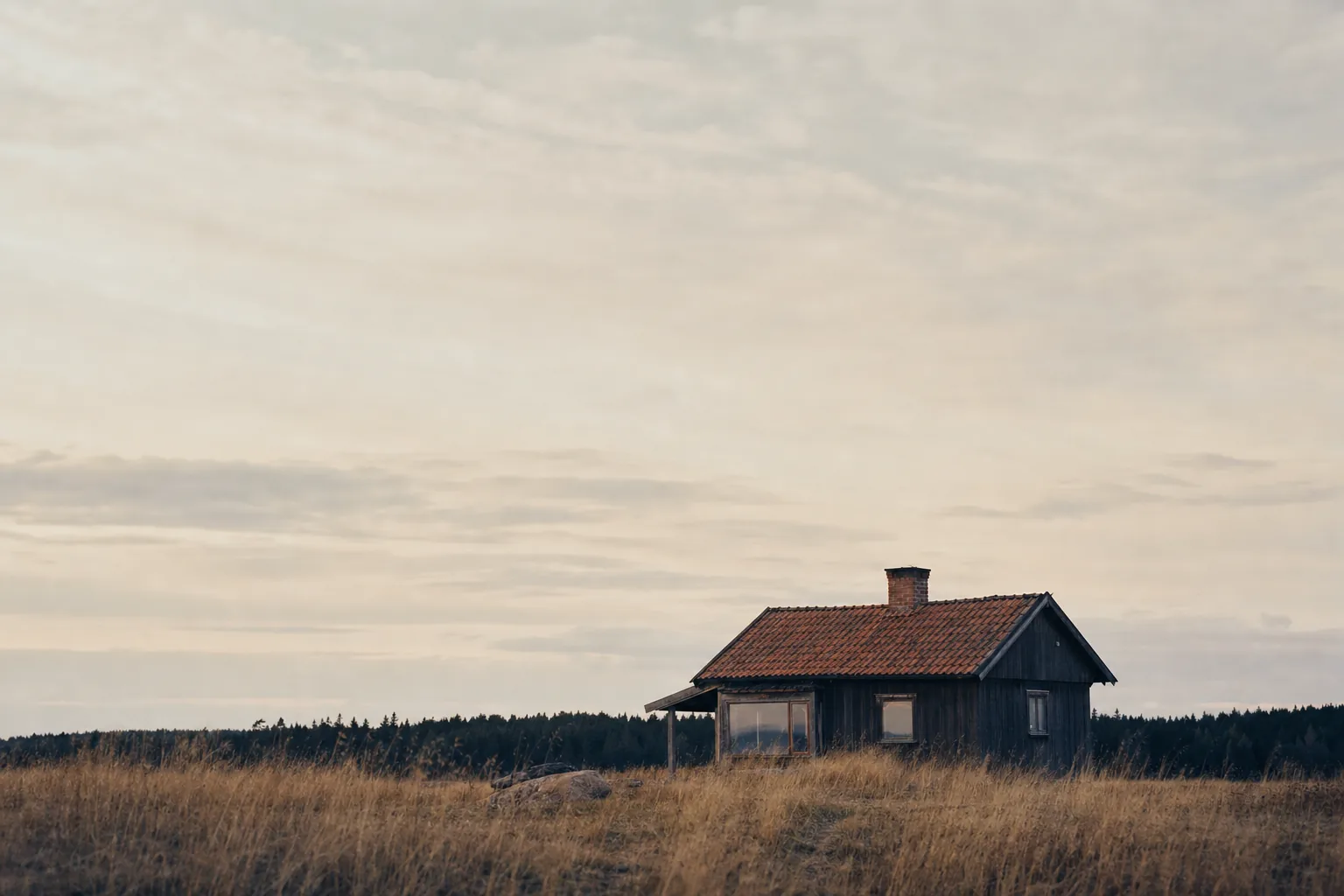 A small single-storey cabin with a red tin roof standing alone in a wide field of dry grass (example photo)